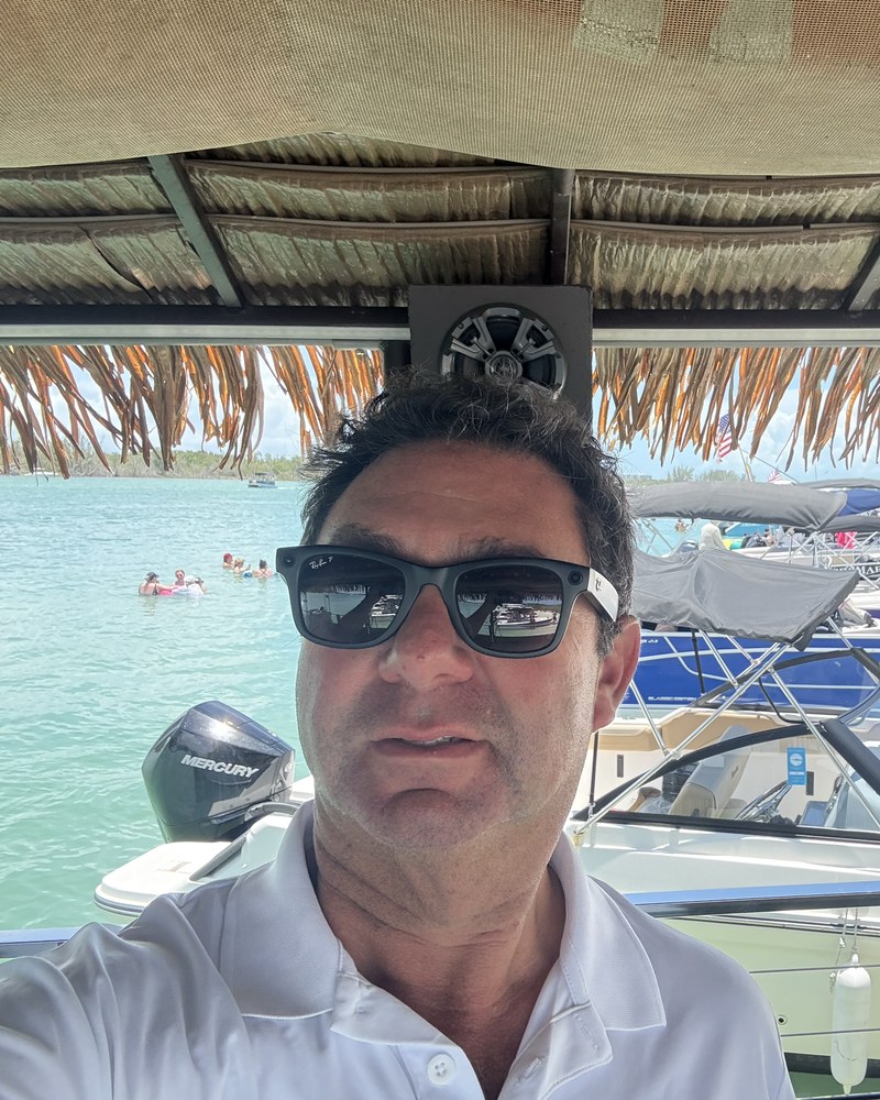 Captain Evan in sunglasses and a white polo under the tiki boat's thatched roof, boats and a beach behind him