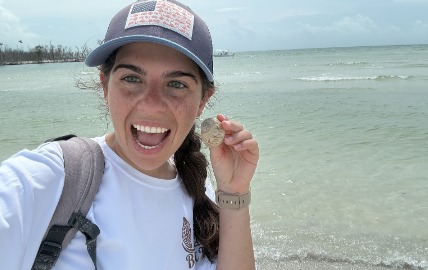 First Mate Taylor in a trucker cap holding up a small shell find on a calm Gulf beach